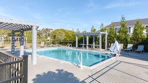 A swimming pool with lounge chairs and a pergola, surrounded by a fence and trees. - Cast A Wave | Steps to Beach & Pool (Corolla)