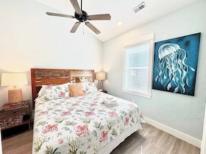 A bedroom with a bed, a ceiling fan, a nightstand, a lamp, a window with blinds, and a wall painting.
