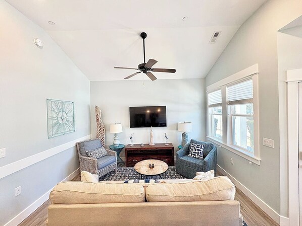 A living room with a ceiling fan, a flat-screen TV, two sofas, a coffee table, and a window with blinds. - Cast A Wave | Steps to Beach & Pool (Corolla)