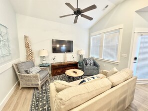 Una sala de estar con ventilador de techo, televisor de pantalla plana, sofá, sillón, mesa de centro y alfombra estampada.