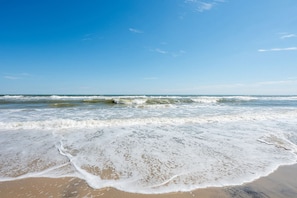 A beach with waves approaching the shore, clear skies, and a calm sea. - Cast A Wave | Steps to Beach & Pool (Corolla)
