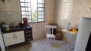 Una pequeña cocina con estufa a gas, un lavaplatos y una ventana. Hay una silla y una mesa con diversos objetos encima.