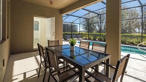 A covered patio with a glass-top table and chairs, a ceiling fan, and a view of a swimming pool and greenery. - Sun-soaked At Solterra (Davenport)