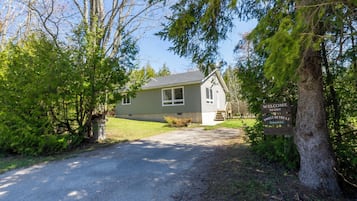 Een huis met een grindpad dat leidt naar een voordeur, omringd door bomen en een bord dat een gastvrij verblijf aankondigt.