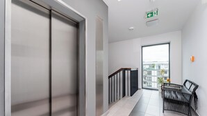A modern hallway with a sliding door, a black wicker bench, a railing, and a large window with a view of buildings.