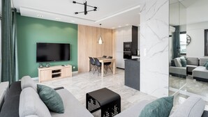 A modern living room with a flat-screen TV, a dining area with a wooden table and chairs, and a kitchen in the background.
