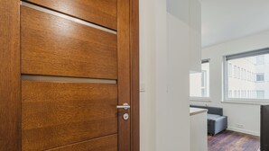 A wooden door with a modern handle, a hallway with a grey bench, and a window with a view of buildings. - Warta River Cozy Vibe Studio (Poznań)