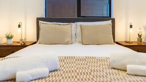 A neatly made bed with two pillows, a checkered rug, and two bedside tables with lamps and small plants.