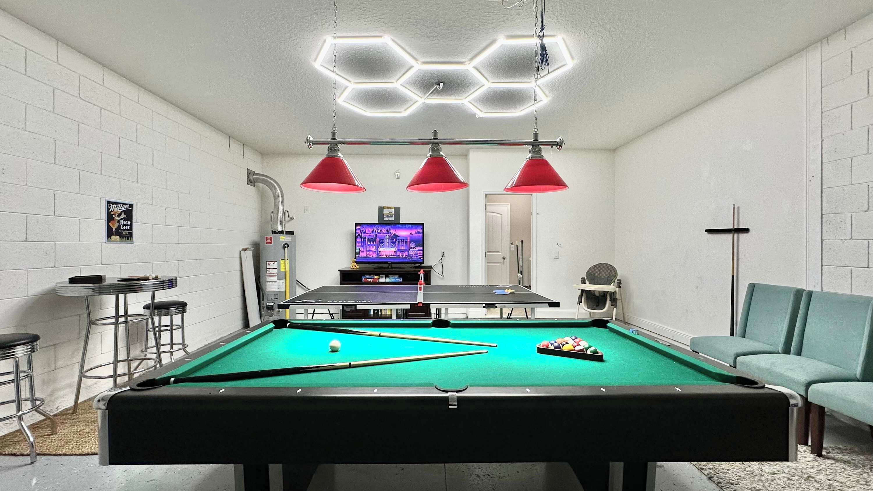 Une table de billard avec un téléviseur en arrière-plan, un coin bar avec des tabourets et un canapé turquoise.