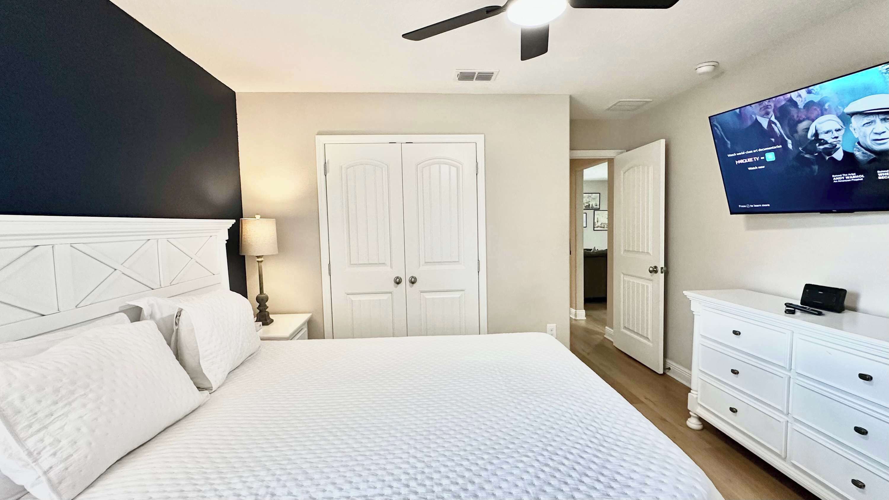 A bedroom with a bed, a TV mounted on the wall, a ceiling fan, and a door leading to another room.