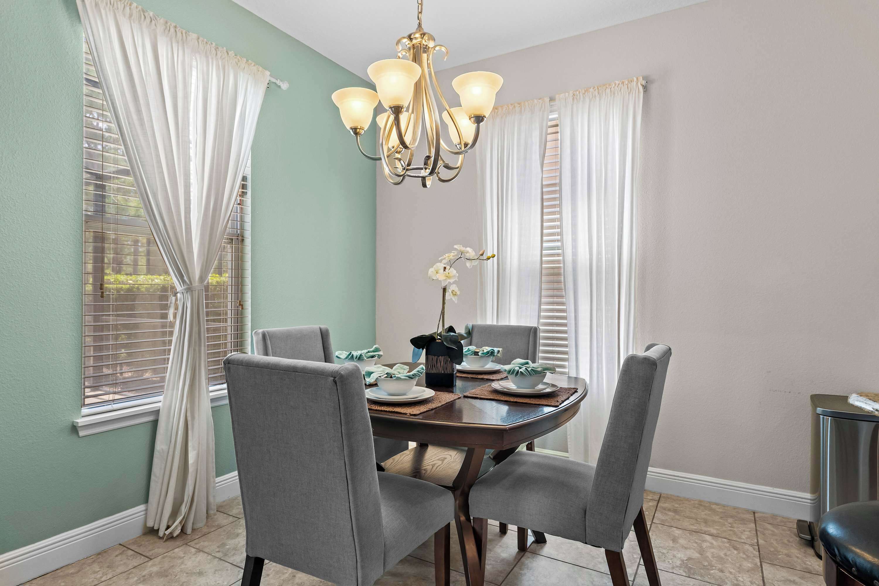 A dining area with a round table, grey chairs, a chandelier, and white curtains.