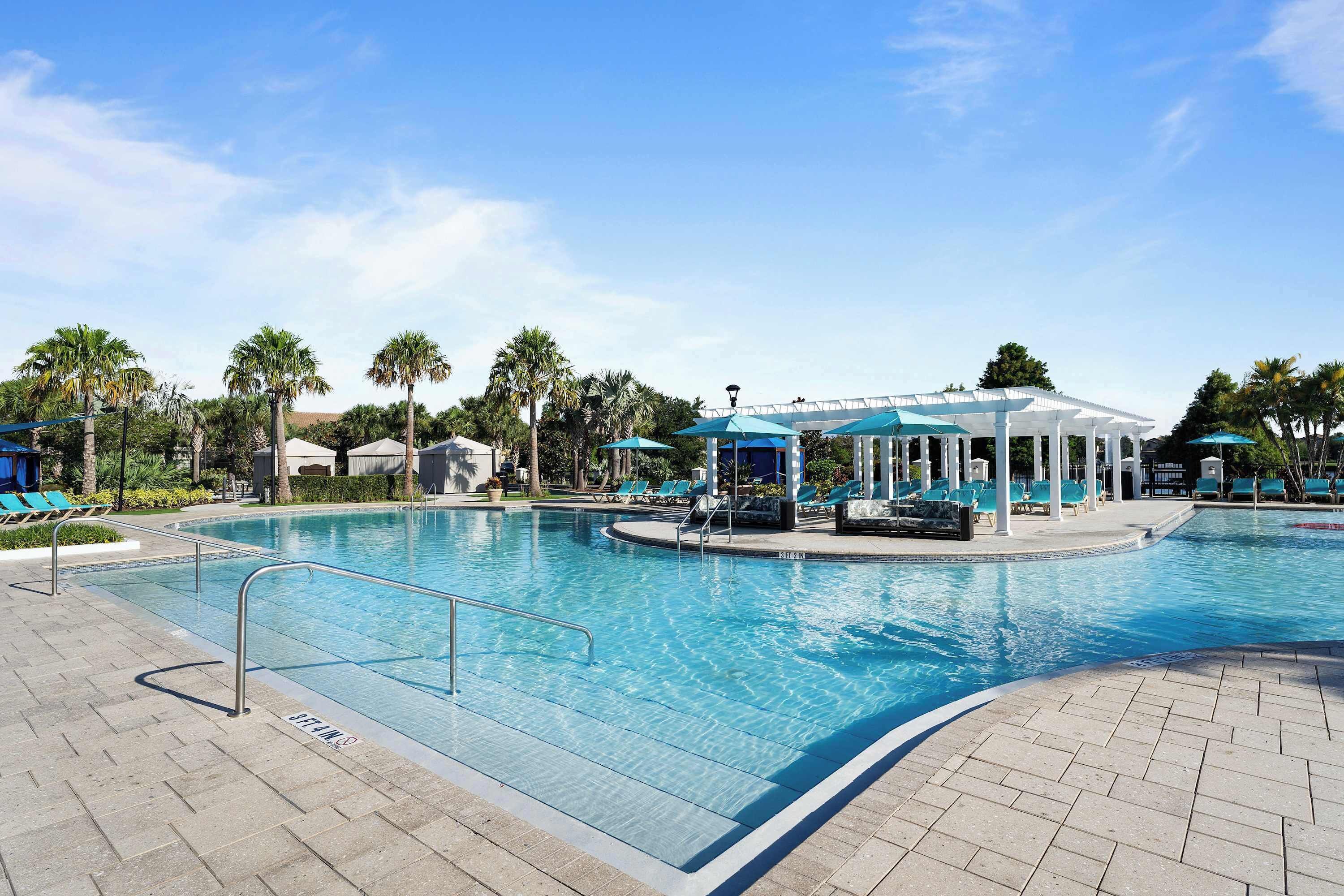 Ein Swimmingpool mit Liegestühlen und Sonnenschirmen, umgeben von Palmen und einem überdachten Sitzbereich.