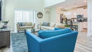 A spacious living room with a large blue sofa, a beige sofa, and a coffee table. There is a kitchen area with modern appliances and a dining area with a table and chairs.