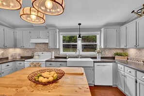 Une cuisine avec des meubles blancs, un îlot central en bois surmonté d’un panier de fruits, une fenêtre offrant une belle vue et des luminaires suspendus.