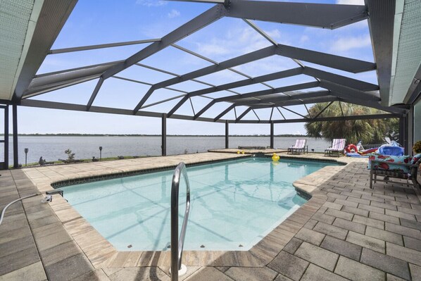 A covered swimming pool with a view of the water body, lounge chairs, and a clear sky.