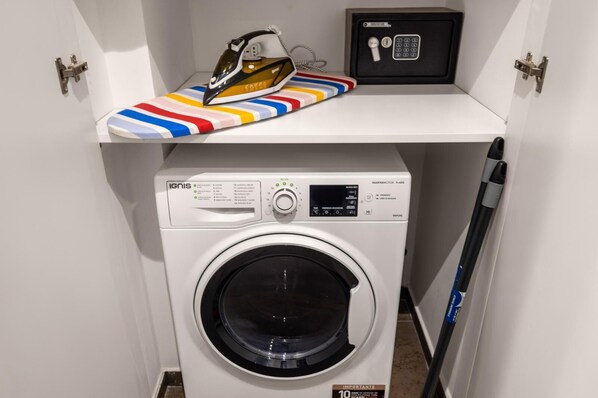 A compact washing machine with a control panel, a safety deposit box, and a colorful ironing board. - Classic One Bedroom Apartment with King Bed (PALERMO)