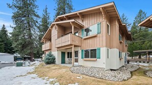 Une maison en bois à deux étages, dotée d’un balcon, d’un porche couvert et d’un petit jardin agrémenté de rochers et de quelques brins d’herbe.