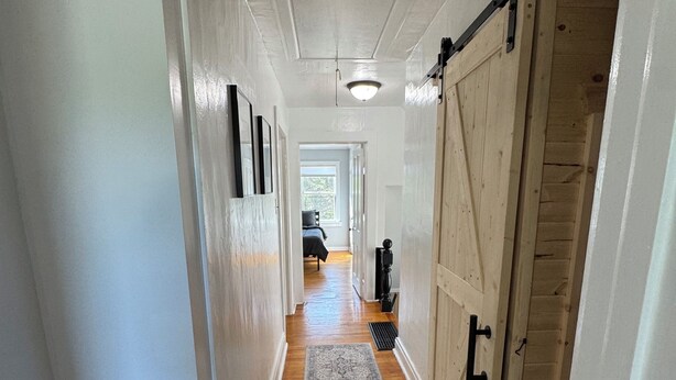 A hallway with wooden flooring, a gray rug, white walls, and a barn-style door. - Charming 3-bedroom 2-bathroom house with ATV access (Calumet)