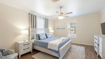 Un dormitorio con cama, mesitas de noche, ventilador de techo, ventana con persianas y vistas al exterior.