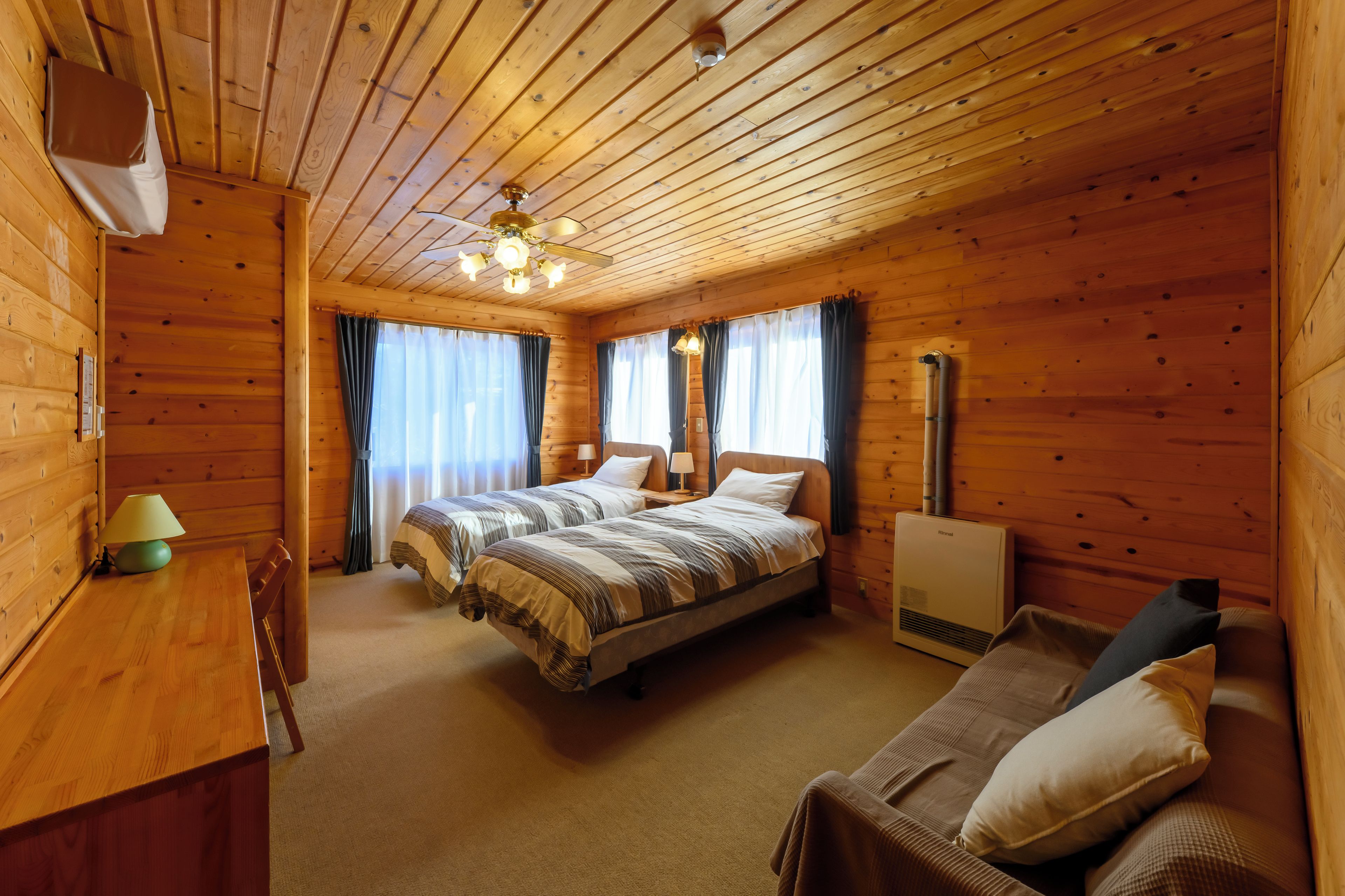 A wooden cabin room with two beds, a desk, a chair, a heater, and a ceiling fan.