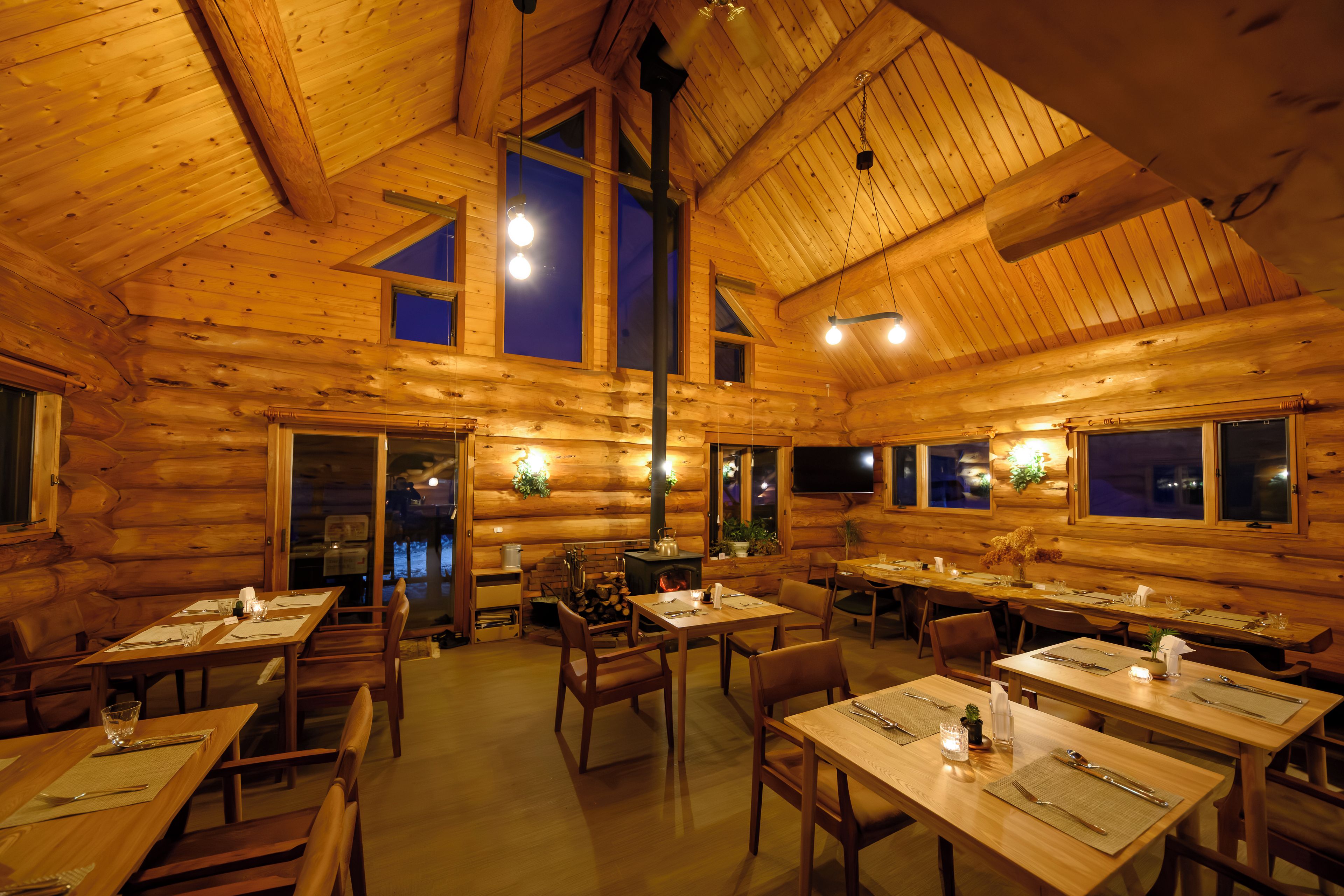 A log cabin interior with multiple dining tables and chairs, a fireplace, and large windows.