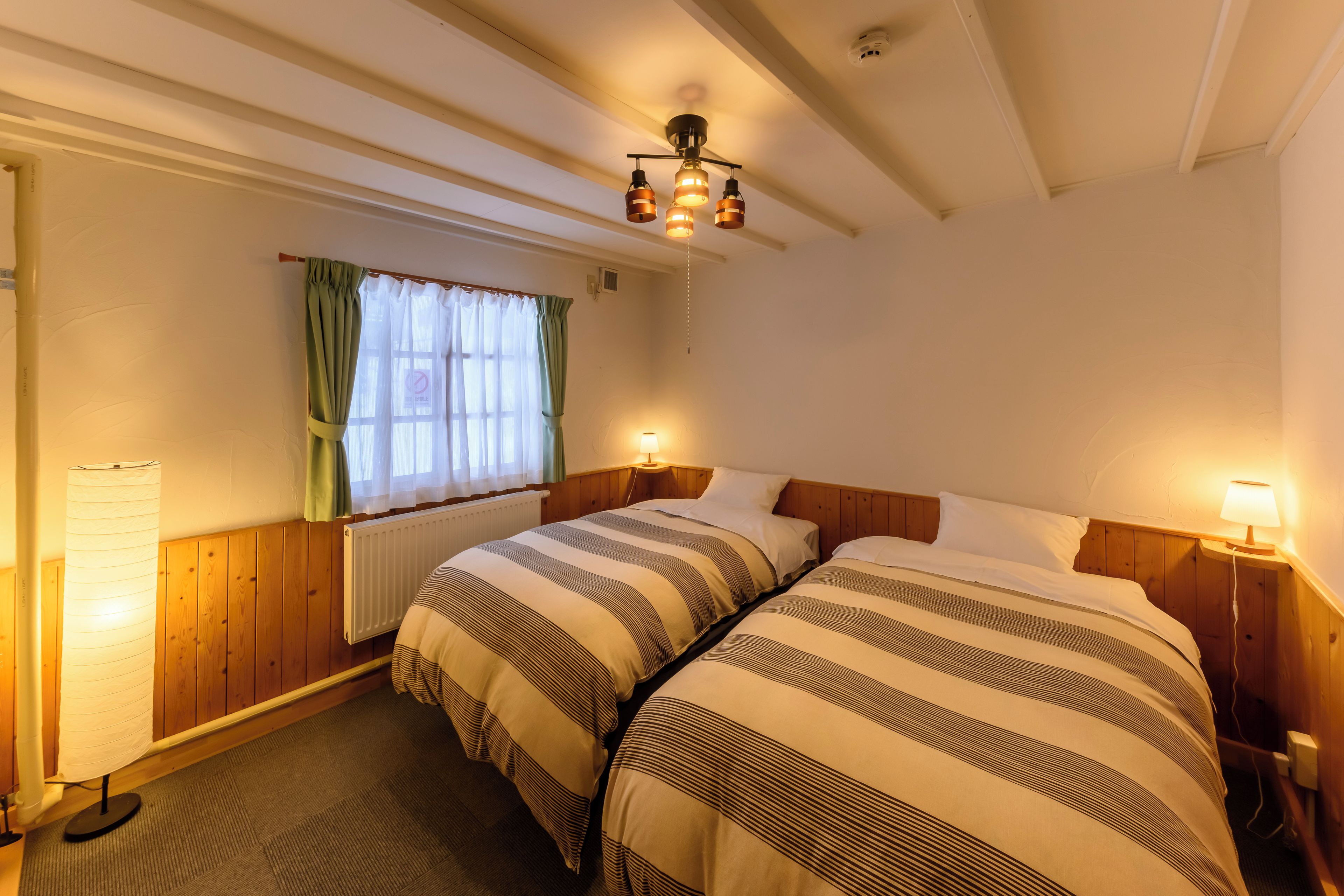 A room with two beds, a window with curtains, a radiator, and a ceiling light fixture.