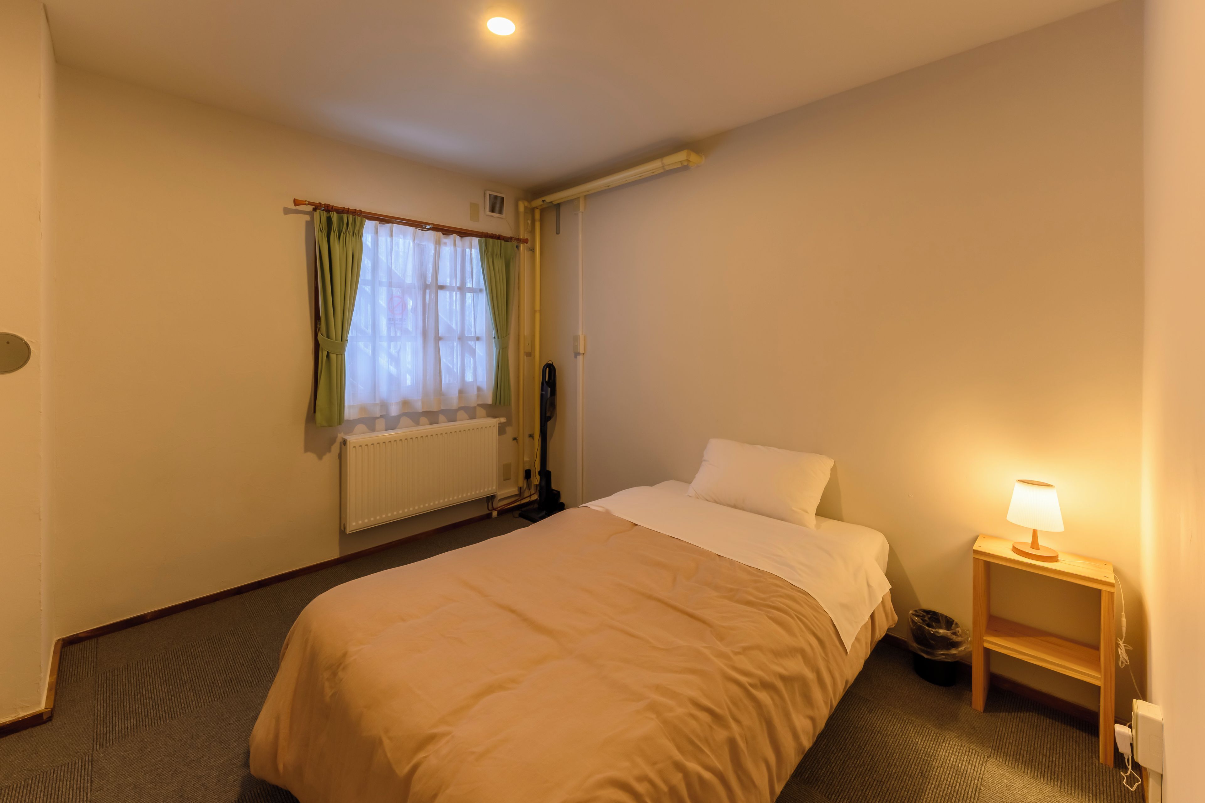 Une chambre d’hôtel avec un lit, une table de chevet avec une lampe, un radiateur et une fenêtre avec des rideaux.
