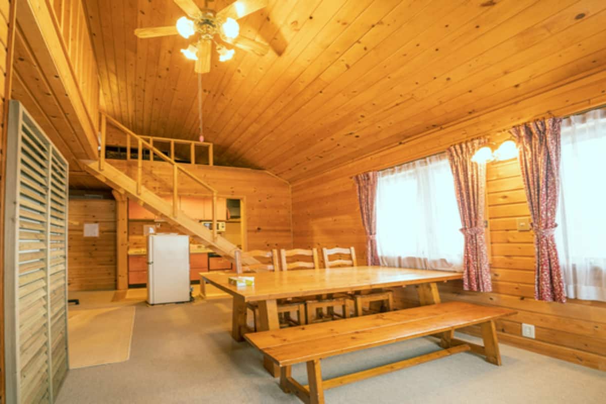 A wooden cabin interior with a dining table, benches, a staircase, and a ceiling fan.