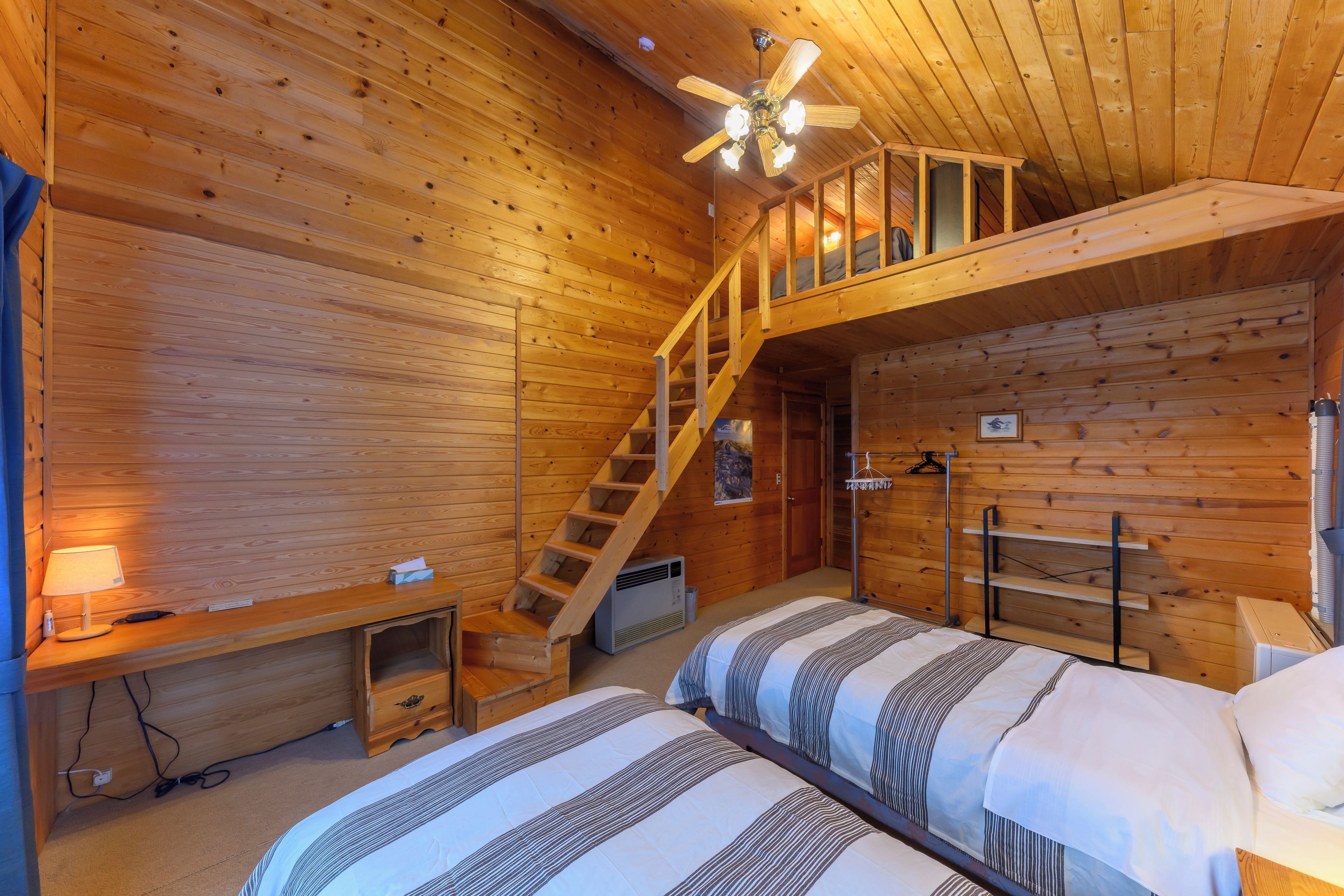 A wooden cabin room with two beds, a ceiling fan, a staircase, and a heater.