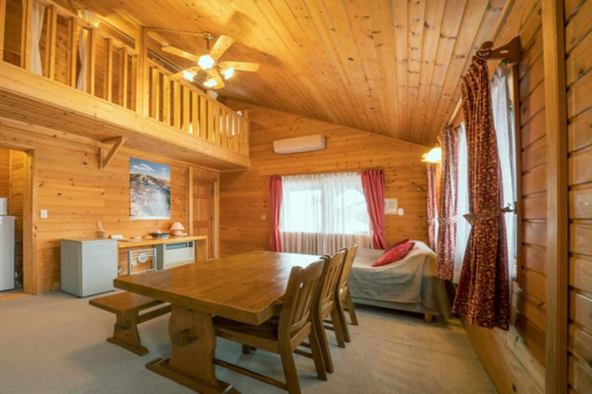 A wooden cabin with a dining area, kitchen, and a bed with curtains.