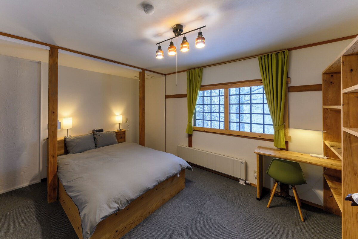 Un dormitorio con cama, lámparas de noche, un escritorio con silla, un radiador y una ventana con cortinas.