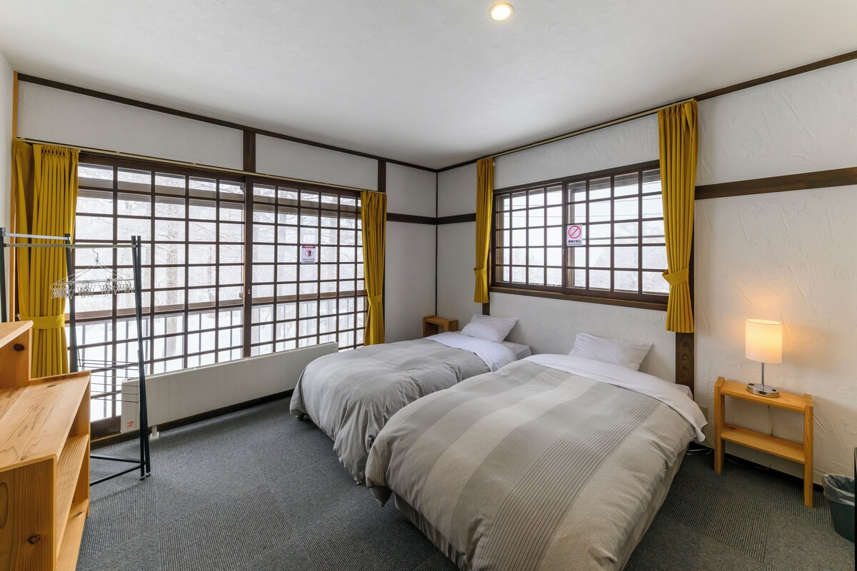 Habitación de hotel con dos camas, un escritorio de madera, una mesita de noche con lámpara y ventanas amplias con cortinas amarillas.