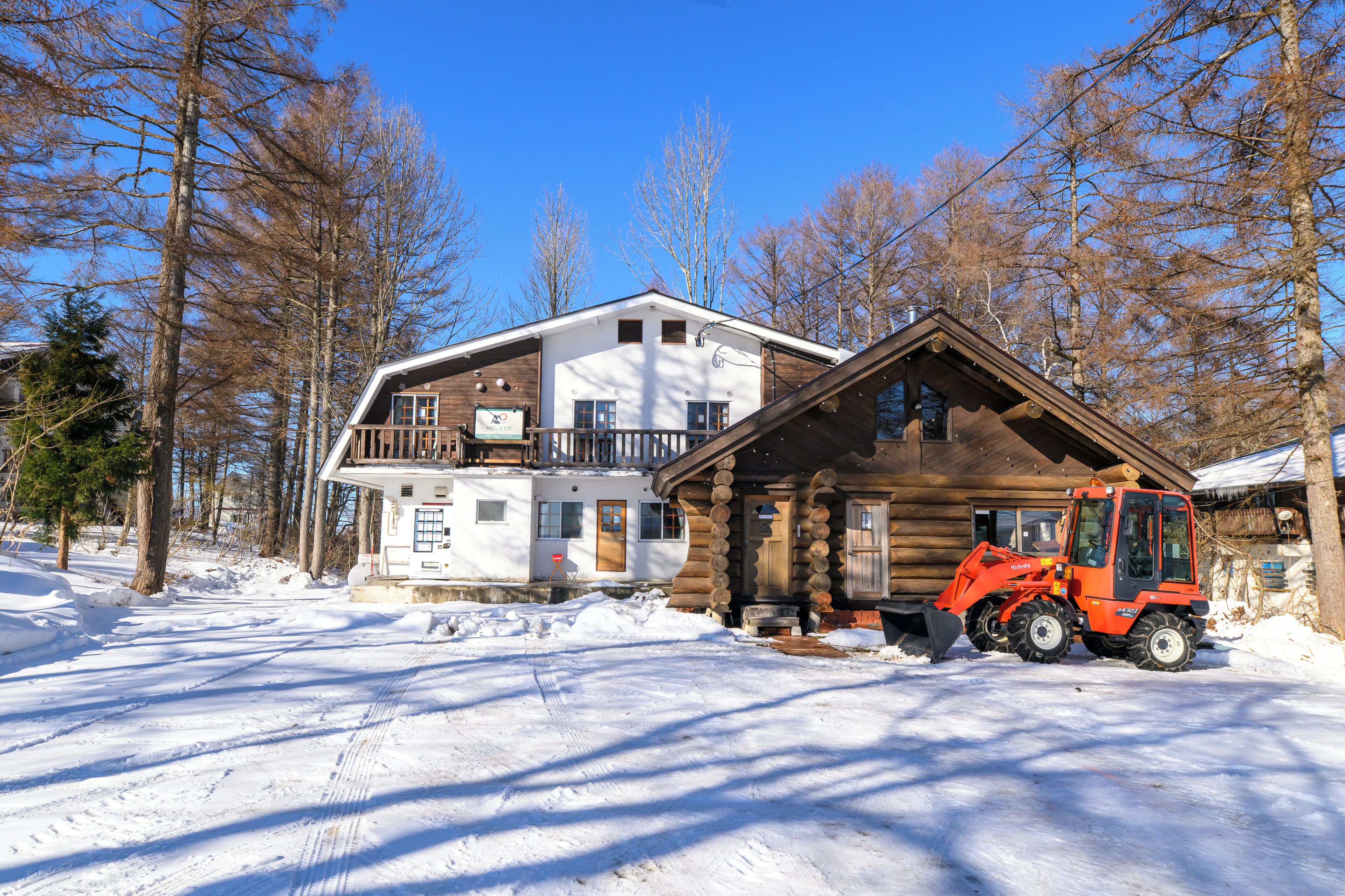 A log cabin with a snow-covered ground, a wooden exterior, and a red vehicle parked in front.