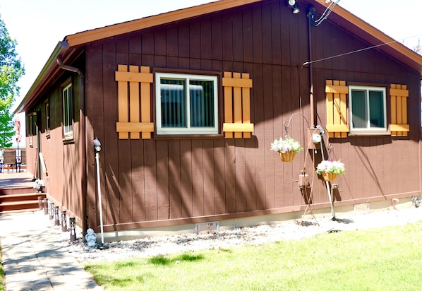 A brown building with two windows, a porch, and a small garden area with potted plants.