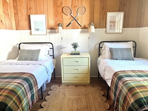 A room with two beds, a nightstand with drawers, a lamp, and wall-mounted tennis rackets.