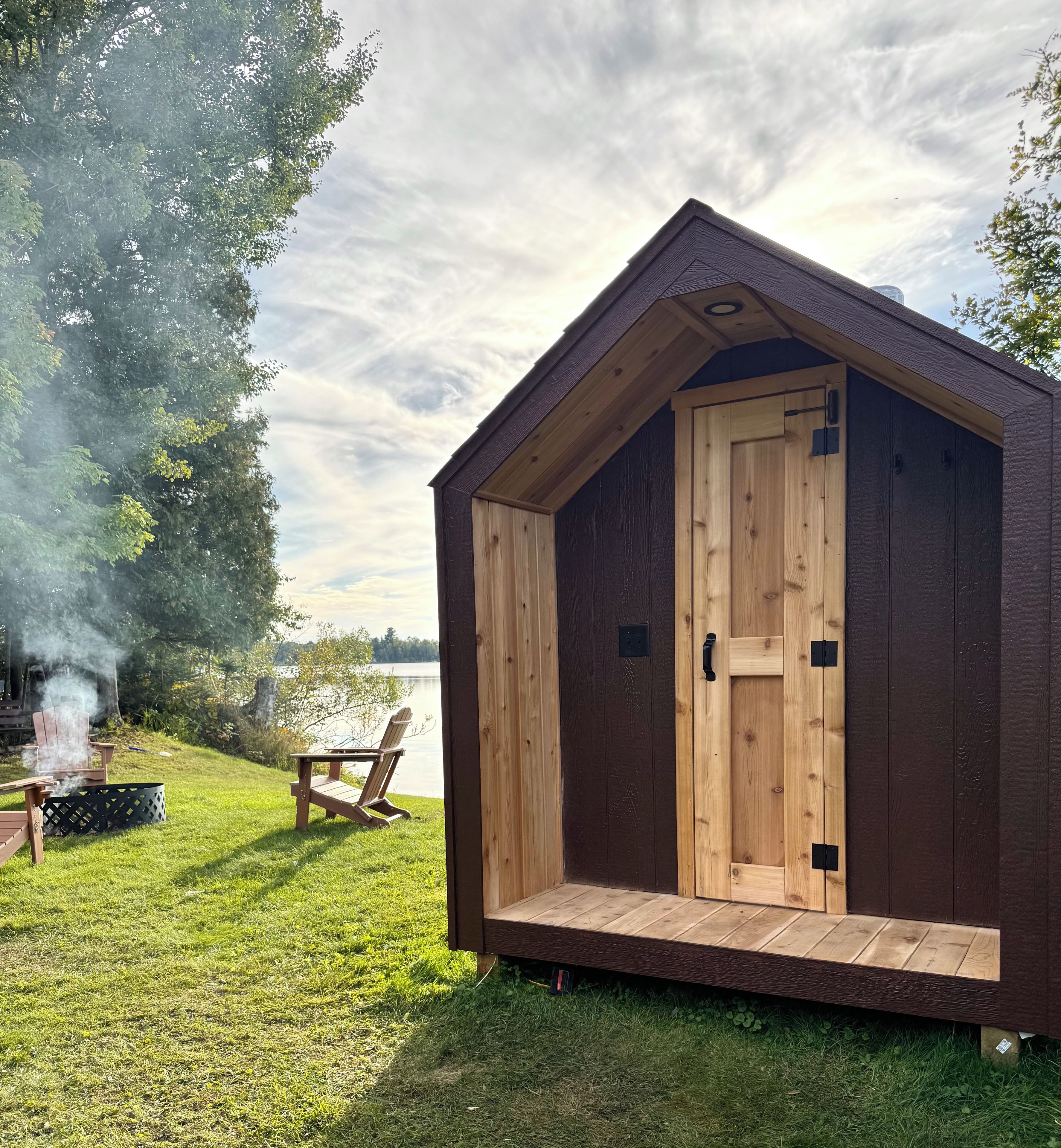 A small wooden cabin with a door and a window, situated on a grassy area with a view of a body of water and trees in the background.