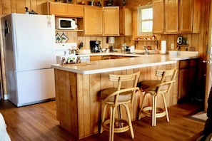 A kitchen with wooden cabinets, a white refrigerator, a microwave, a coffee maker, and two bar stools.