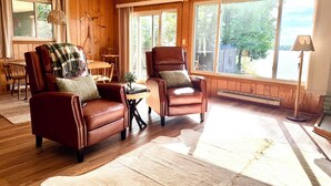 A cozy living room with leather armchairs, a wooden dining table, and large windows offering a view of the outdoors.