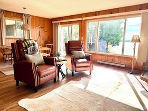 A cozy living room with leather armchairs, a wooden dining table, and large windows offering a view of the outdoors.