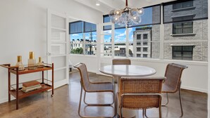 A dining area with a round table and wicker chairs, a wooden side table with books and decorative items, and a view of buildings through large windows. - Luxury Downtown Retreat | Heated Pool Access (Savannah)