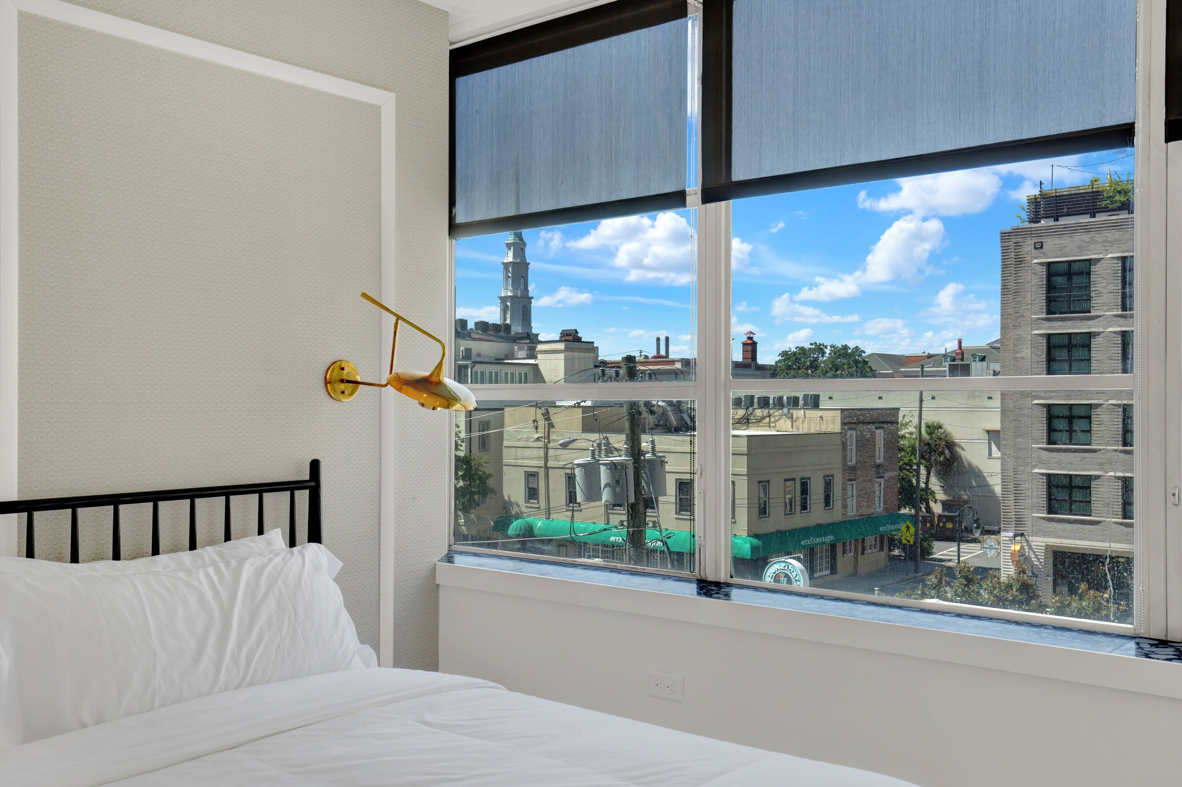 Ein Schlafzimmer mit Bett, Fenster mit Blick auf Gebäude und einer Wandleuchte.