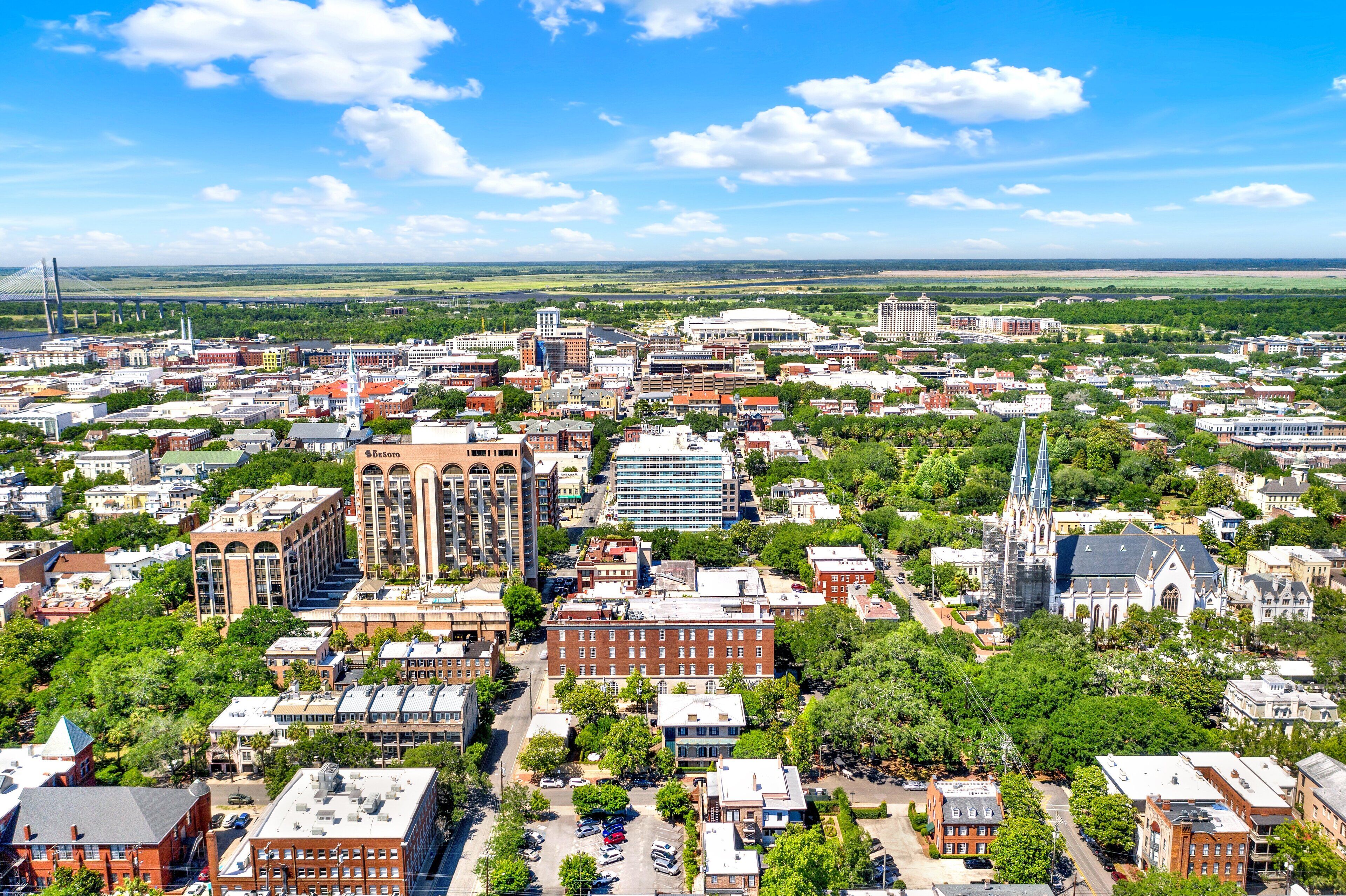 An aerial view of a cityscape with various buildings, including a prominent church with a spire, surrounded by green spaces and a large bridge in the background.
