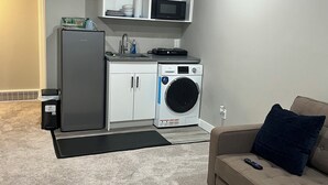 A compact living space with a kitchenette, including a refrigerator, microwave, and washing machine. - RaphaSuites (Regina)