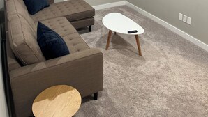 A modern living room with a sectional sofa, a small round coffee table, and a flat-screen TV mounted on the wall. - RaphaSuites (Regina)