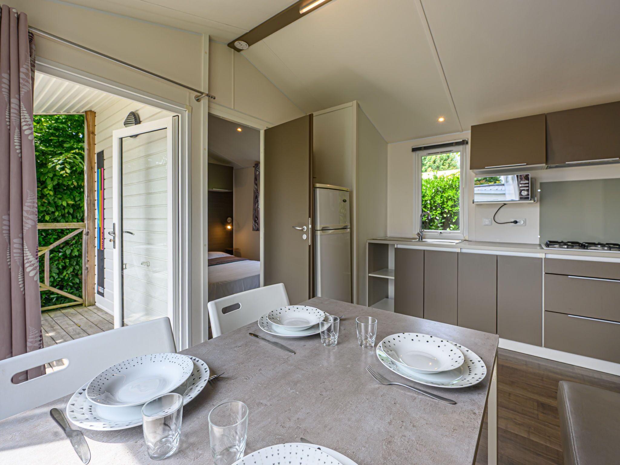 A modern kitchen with a dining area, including a table set for four, a refrigerator, and a window with a view of greenery.