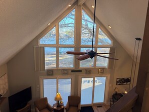 A high-ceiling room with large windows, a ceiling fan, and a TV on a wooden stand.