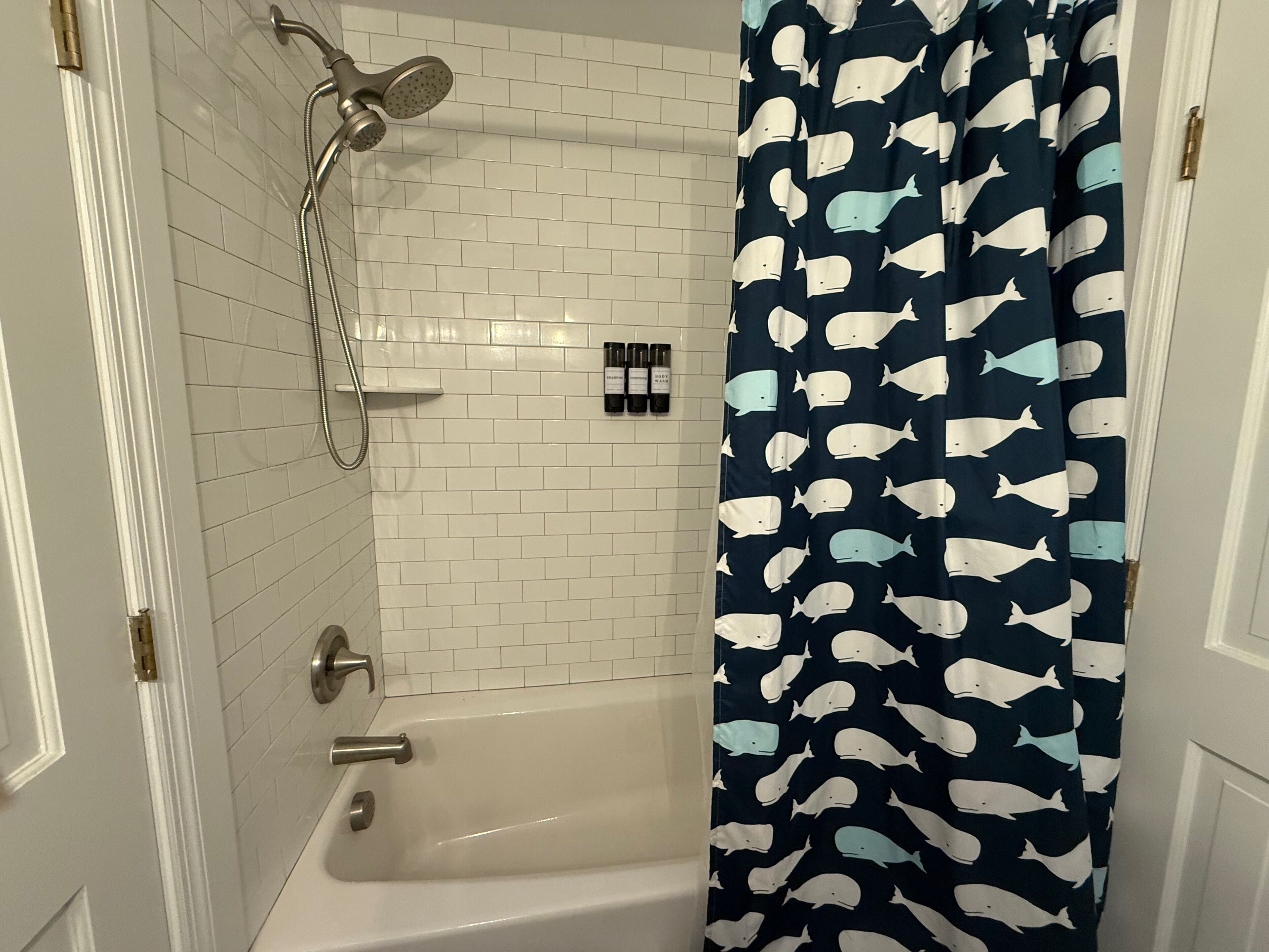 Ein Badezimmer mit weiß gefliesten Wänden, einer weißen Badewanne und einer Dusche inklusive Duschbrause. Ein Vorhang mit Walmotiv ziert das Zimmer.