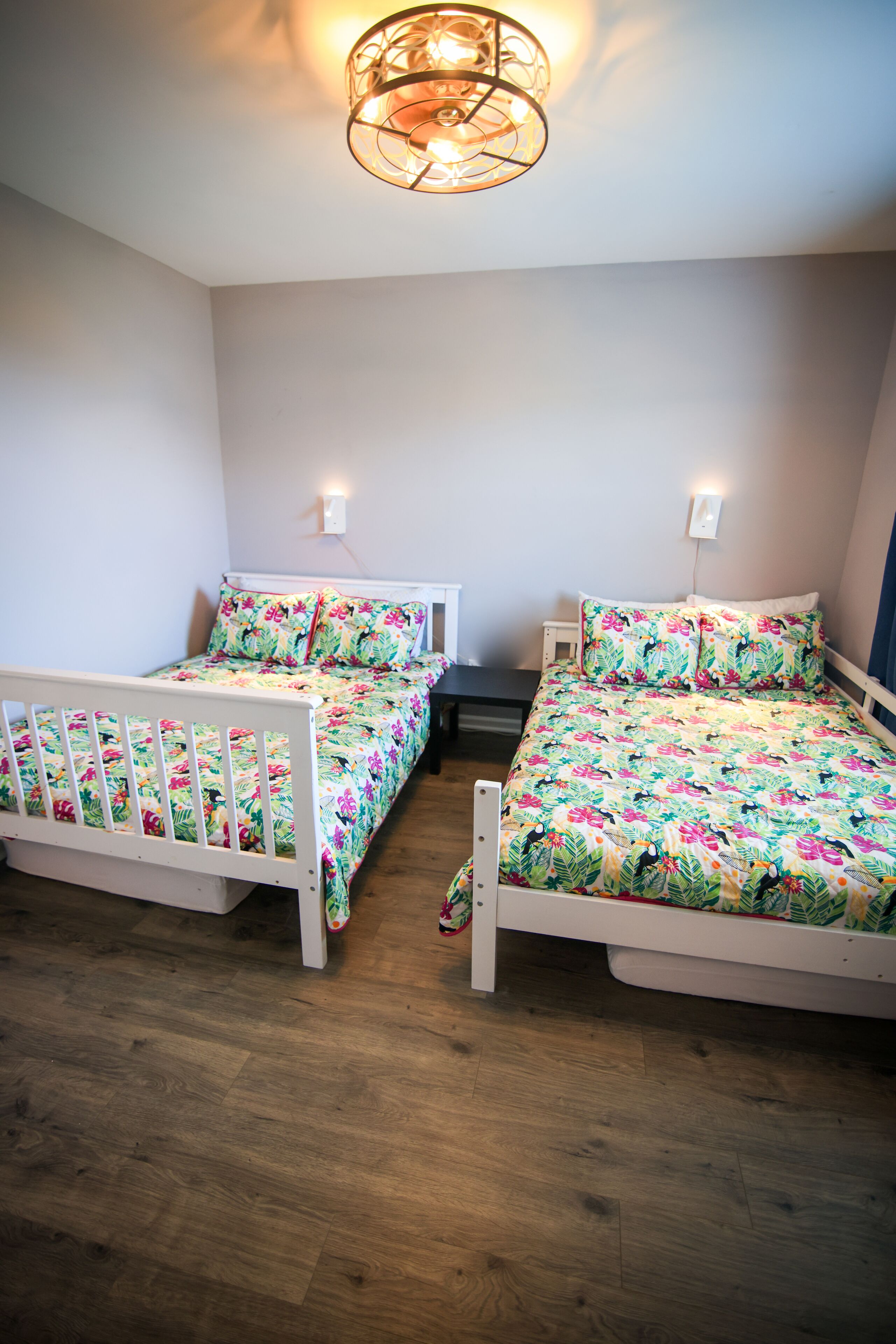 Une chambre avec deux lits simples, chacun agrémenté d’une couvre-lit à motifs floraux et d’une tête de lit blanche. Un luminaire est fixé au plafond, au-dessus des lits.
