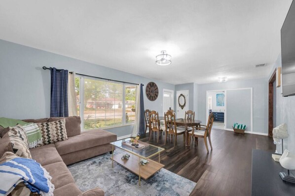 A living room with a sofa, coffee table, and dining table with chairs. There is a large window, a clock on the wall, and a door leading to another room.