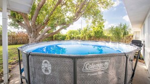 Una piscina exterior con una cubierta de la marca Coleman, rodeada de árboles y una parrilla para barbacoa.